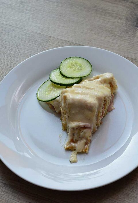 Een bord met een stuk Toast cirkel met ham en kaas, met plakjes komkommer.