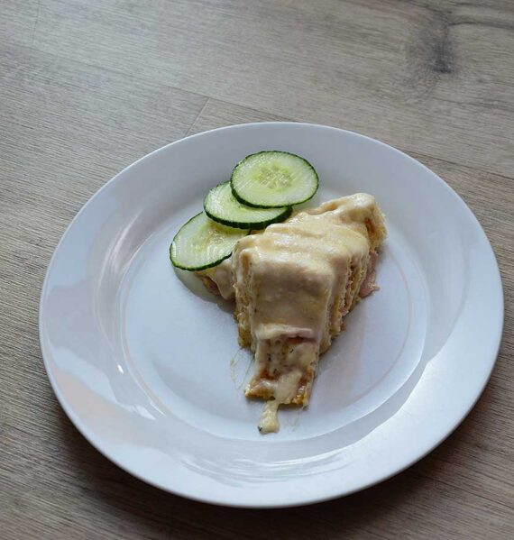 Een bord met een stuk Toast cirkel met ham en kaas, met plakjes komkommer.